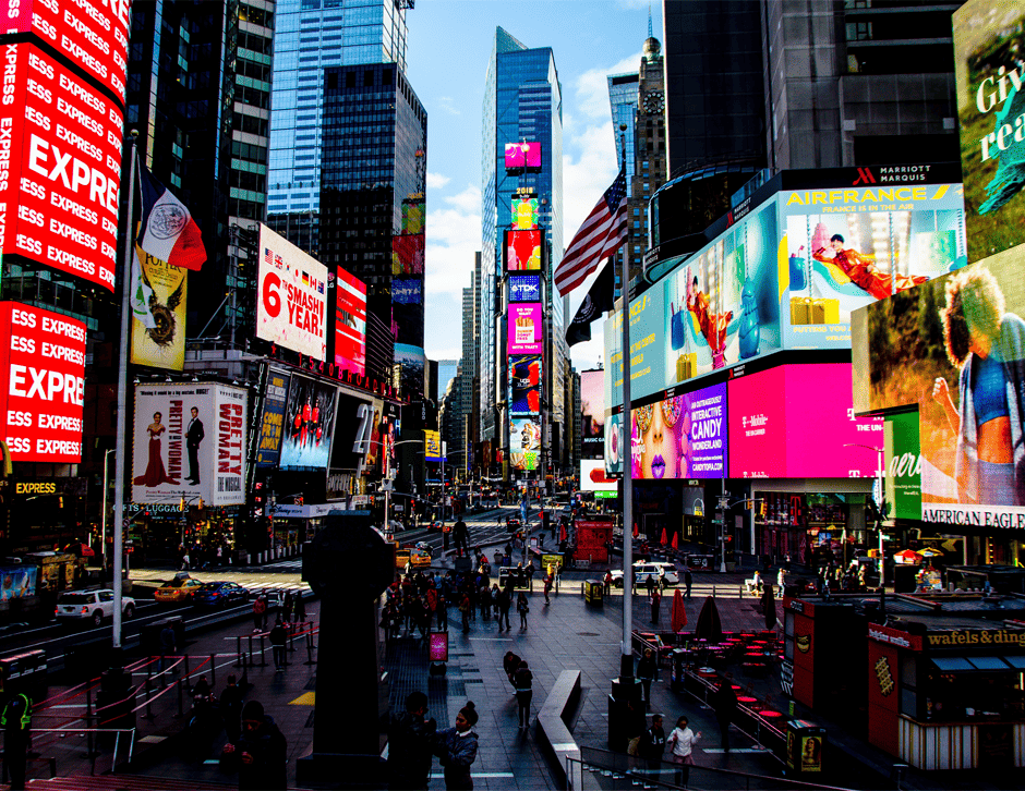 Una fotografía de Times Square, Estados Unidos, donde se muestra un día común. Se ven anuncios espectaculares de productos, descuentos, películas y gente caminando, además de algunos automóviles.
