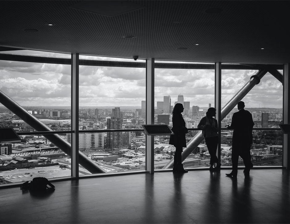 Tres personas paradas platicando en una oficina con una vista a la ciudad preciosa. La imagen esta en blanco y negro.