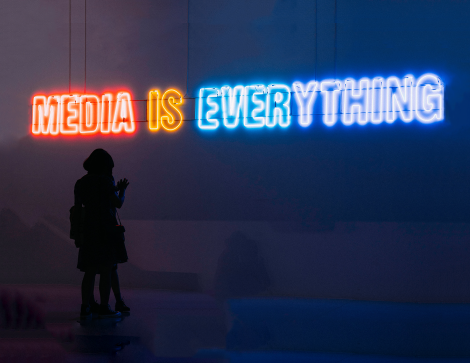 En la imagen se puede ver un letrero en inglés que dice: Media is everything. Con unas personas viendo su celular.